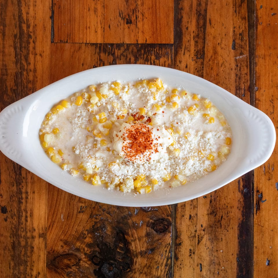 Creamy corn dish topped with crumbled cheese, sour cream, and chili seasoning on a white oval plate atop a wooden table.