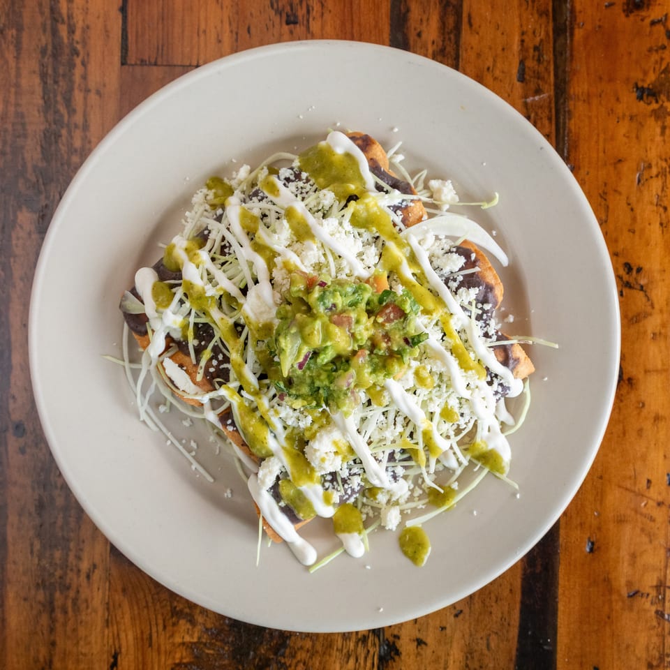 Overhead view of Mexican tostadas topped with black beans, shredded lettuce, cheese, guacamole, crema, and green salsa on a white plate.