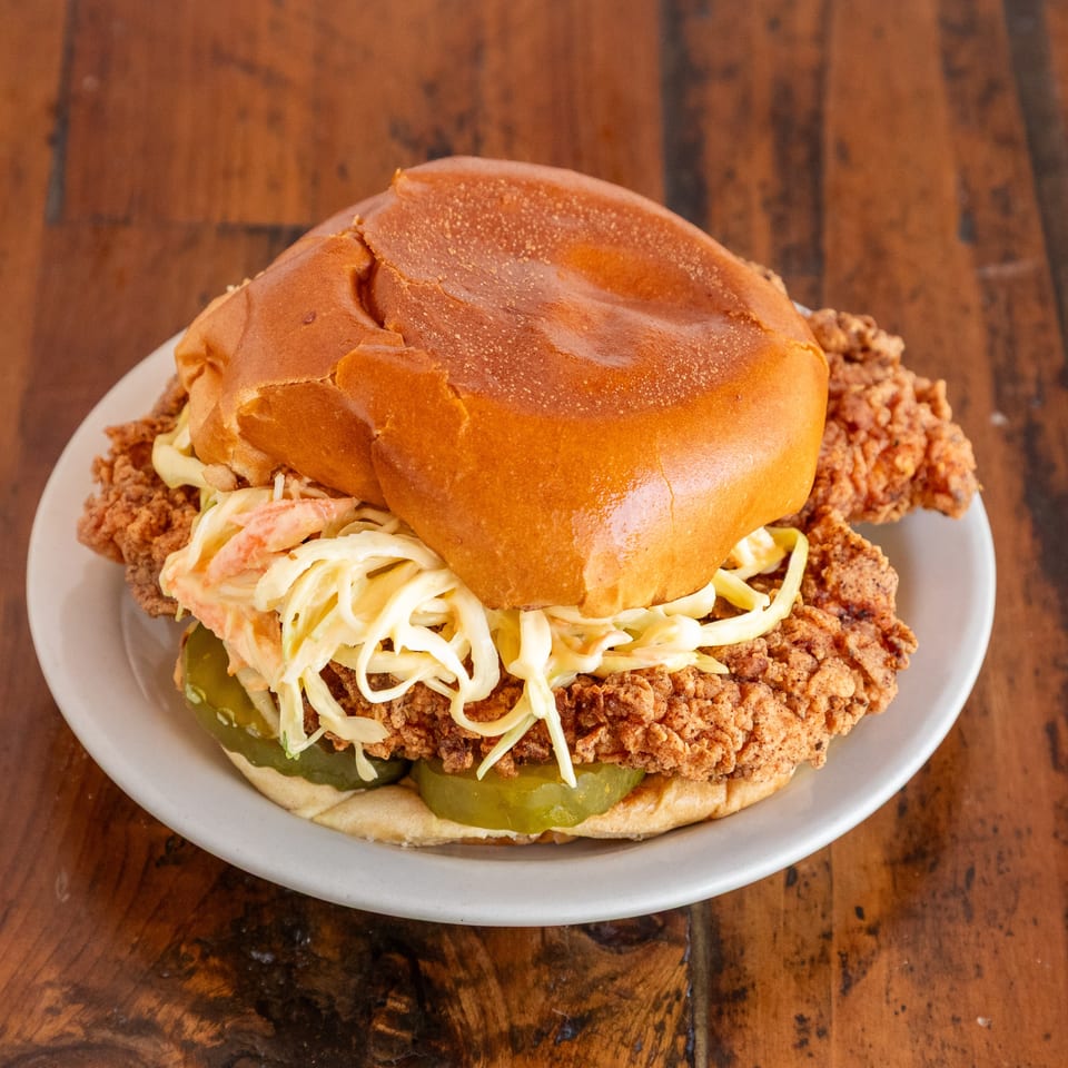Crispy fried chicken sandwich with creamy coleslaw and pickles on a brioche bun, served on a white plate.