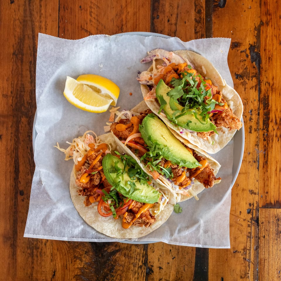 Overhead view of crispy fish tacos topped with avocado, cilantro, slaw, vegetables, and orange sauce, served with a lemon wedge.