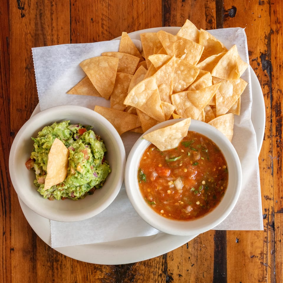 Basket of tortilla chips served with bowls of guacamole and fresh salsa on a wooden restaurant table.