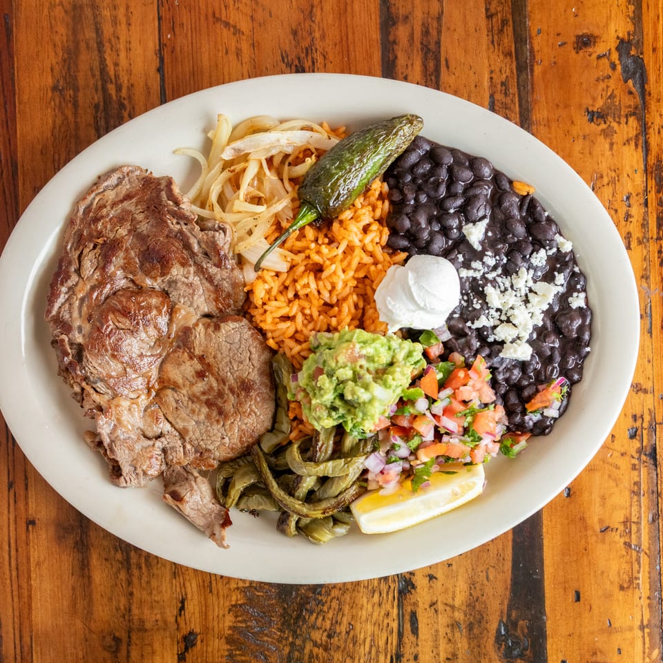 Mexican platter with grilled steak, rice, black beans, guacamole, pico de gallo, roasted peppers, onions, and lime on a wooden table.