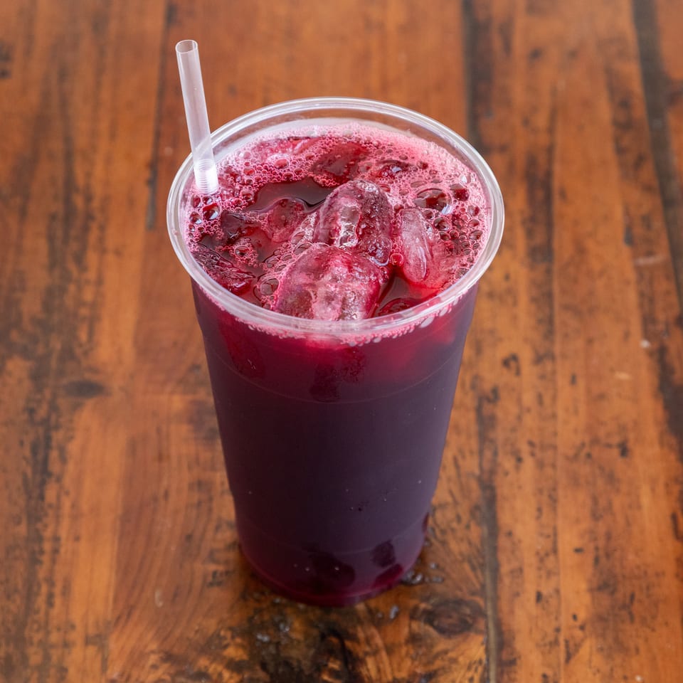 Clear plastic cup of dark red iced beverage with a straw on a rustic wooden table.