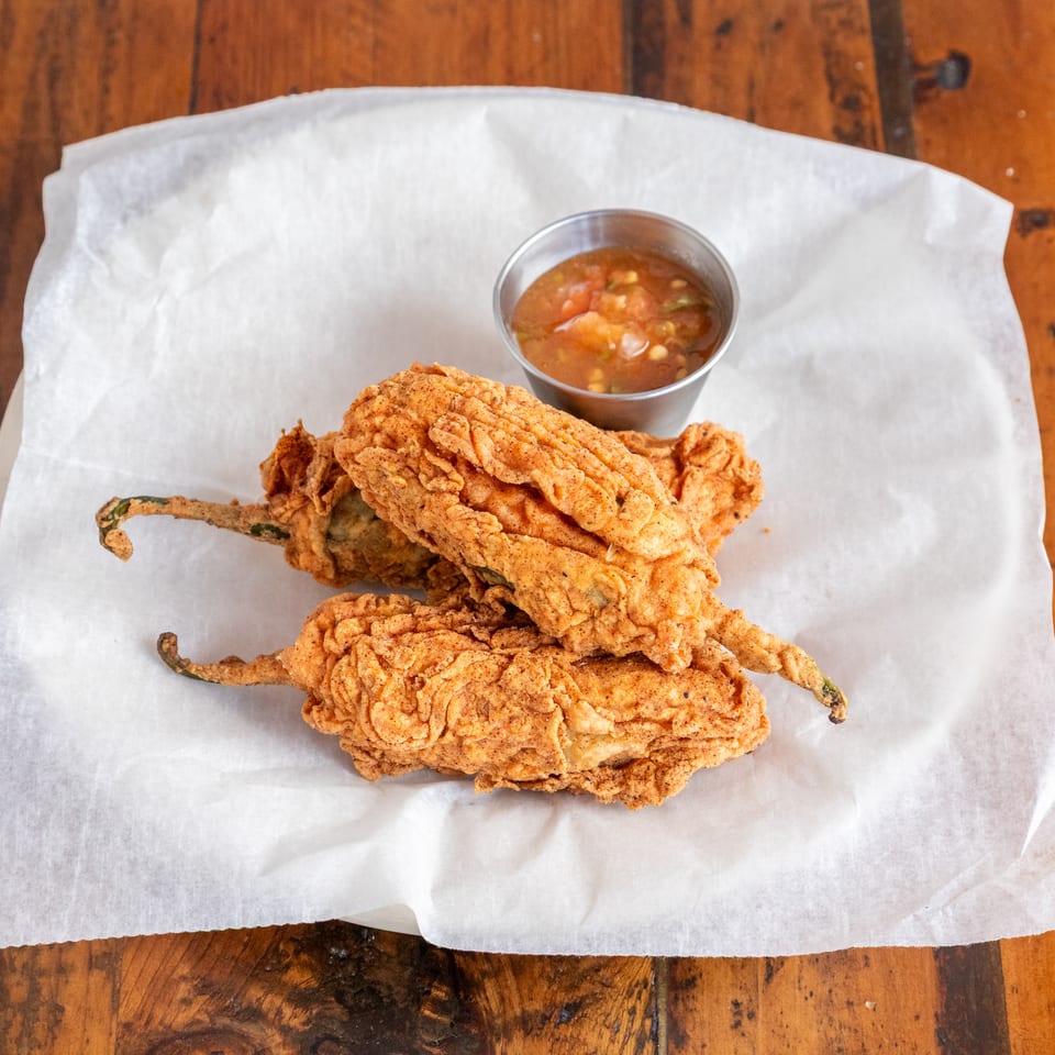 Crispy fried stuffed peppers served on parchment paper with a small bowl of chunky dipping sauce on a wooden table.