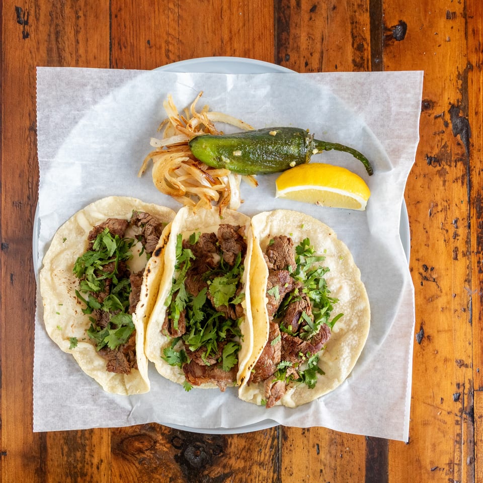 Three steak tacos topped with cilantro, served with grilled onions, a whole jalapeño, and lemon wedge on a wooden table.